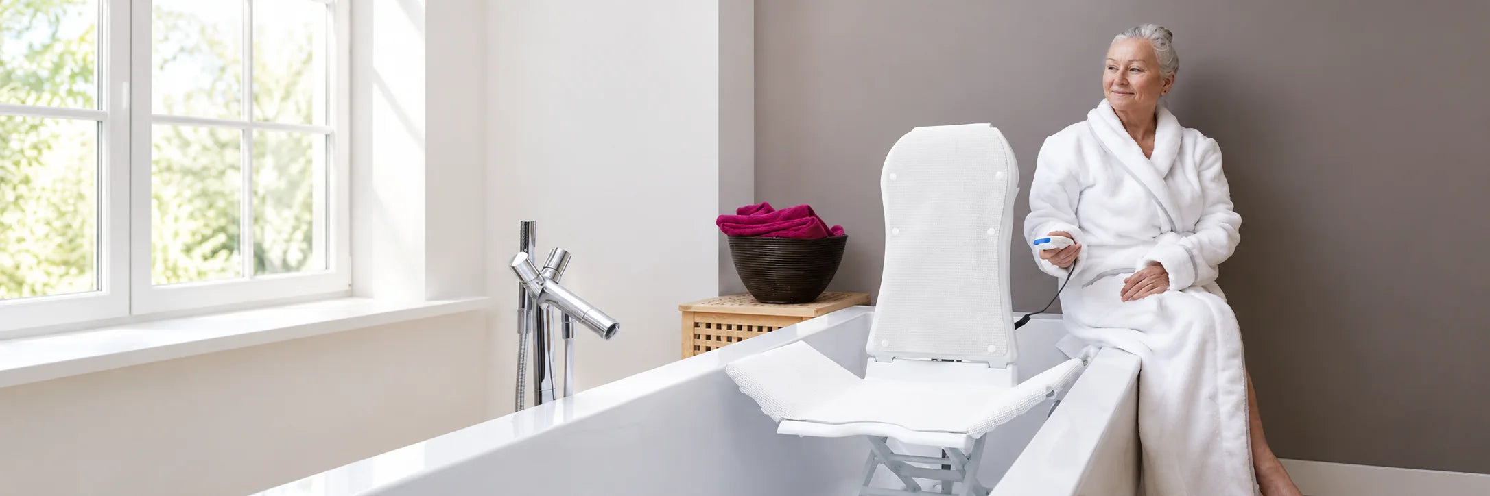 A woman in a white robe sitting on the edge of a bathtub, holding the remote of the bath lift in the tub