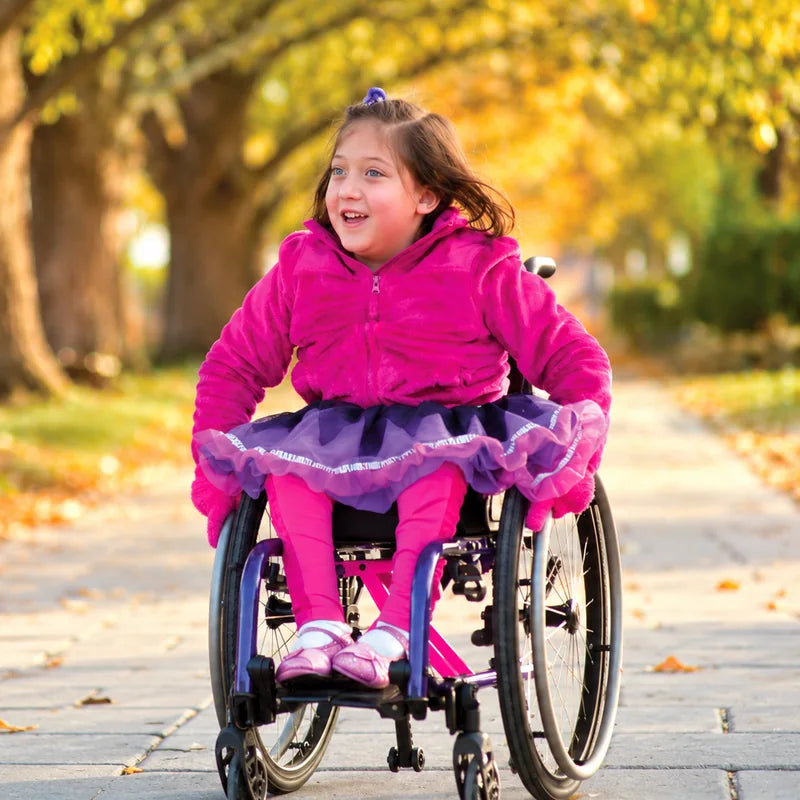 A smiling girl in a pink jacket pushing her own lightweight wheelchair down a tree-lined path in autumn