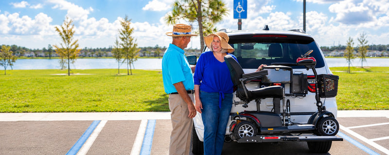 A couple smiling beside their SUV with a mobility scooter on a platform lift at the back