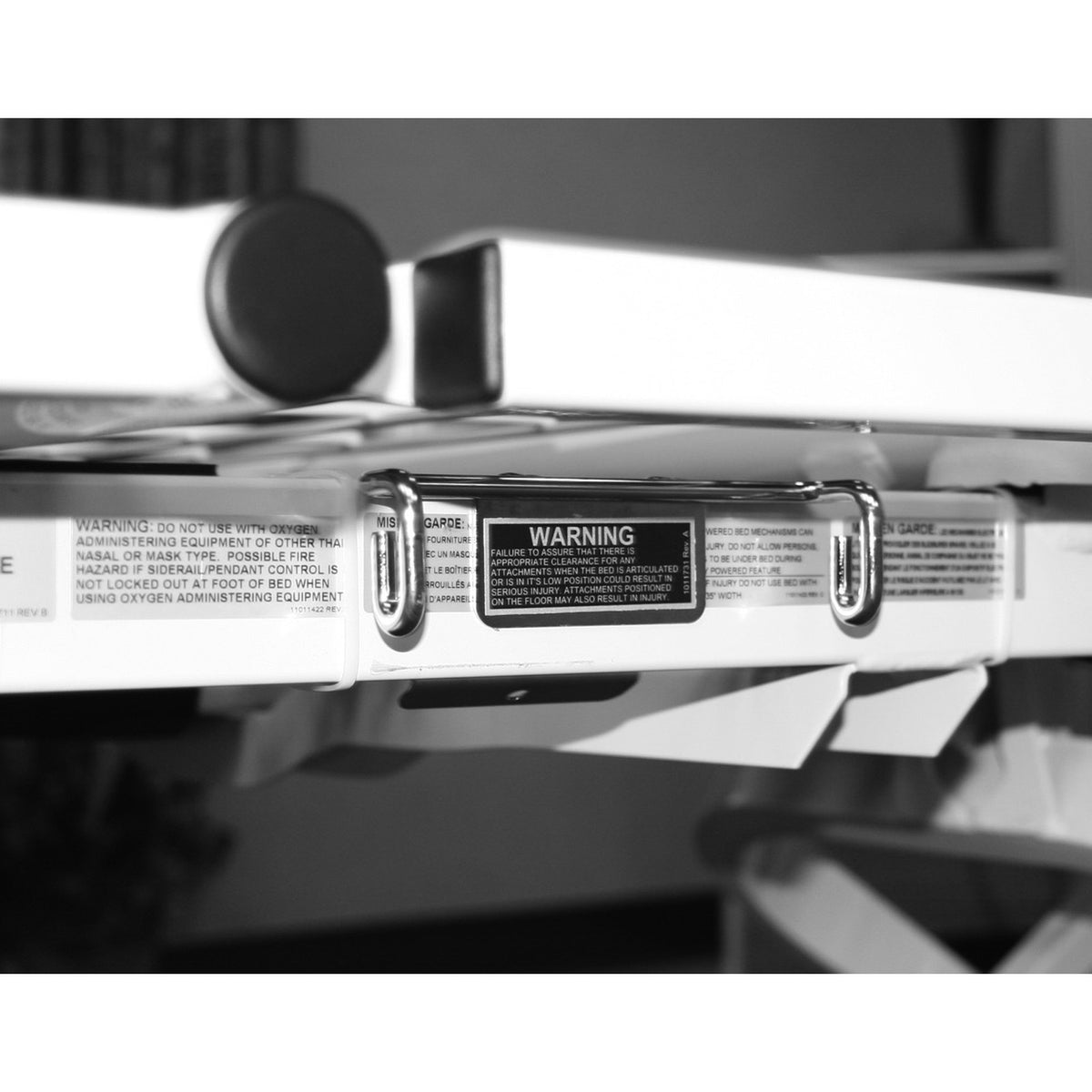 The Joerns drainage bag holder mounted under the edge of a bed frame, with its two hooks