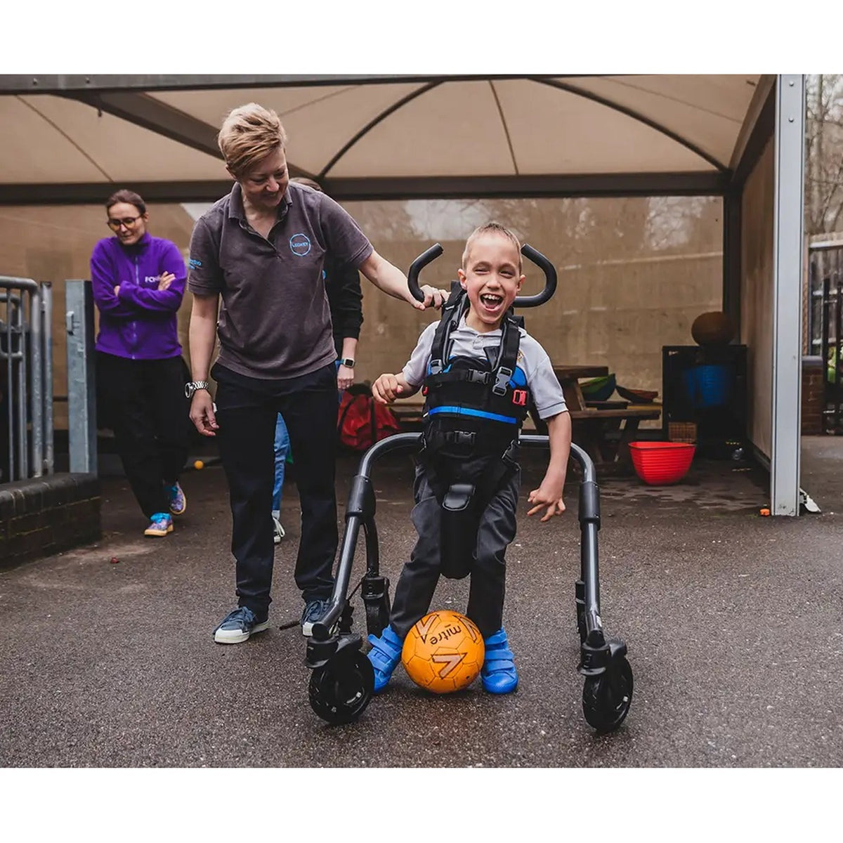 A boy walking upright in the Leckey MyWay+ gait trainer, held by its harness, with the user handles add-on
