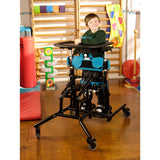 A boy standing at the tray in a Squiggles Stander with blue pads, in a classroom