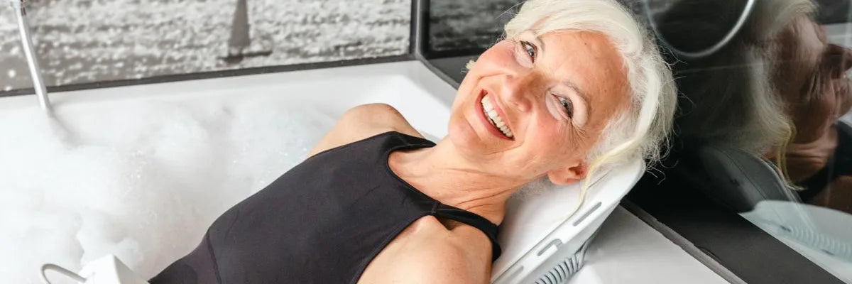 A smiling woman lying back on a bath lift in a bubble bath, holding its remote