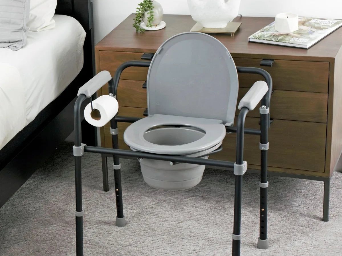 A gray folding commode with padded arms beside a bed