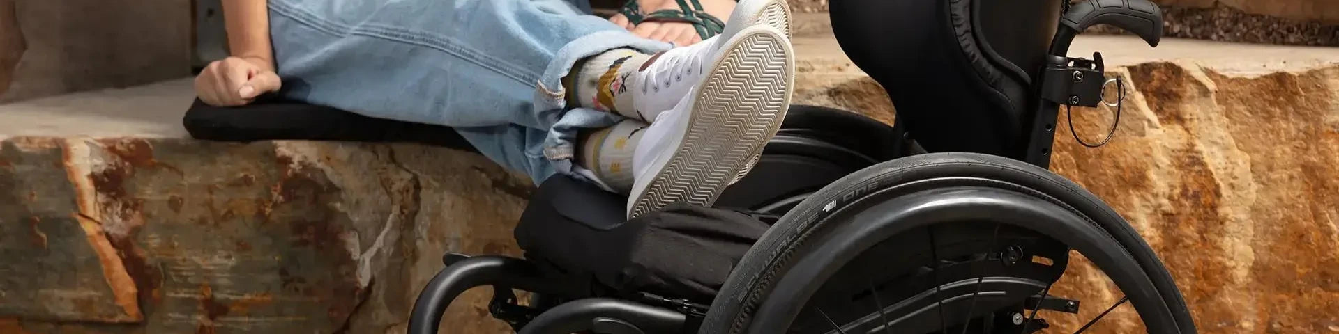 A person sitting beside a black manual wheelchair with a contoured seat cushion and a shaped back support