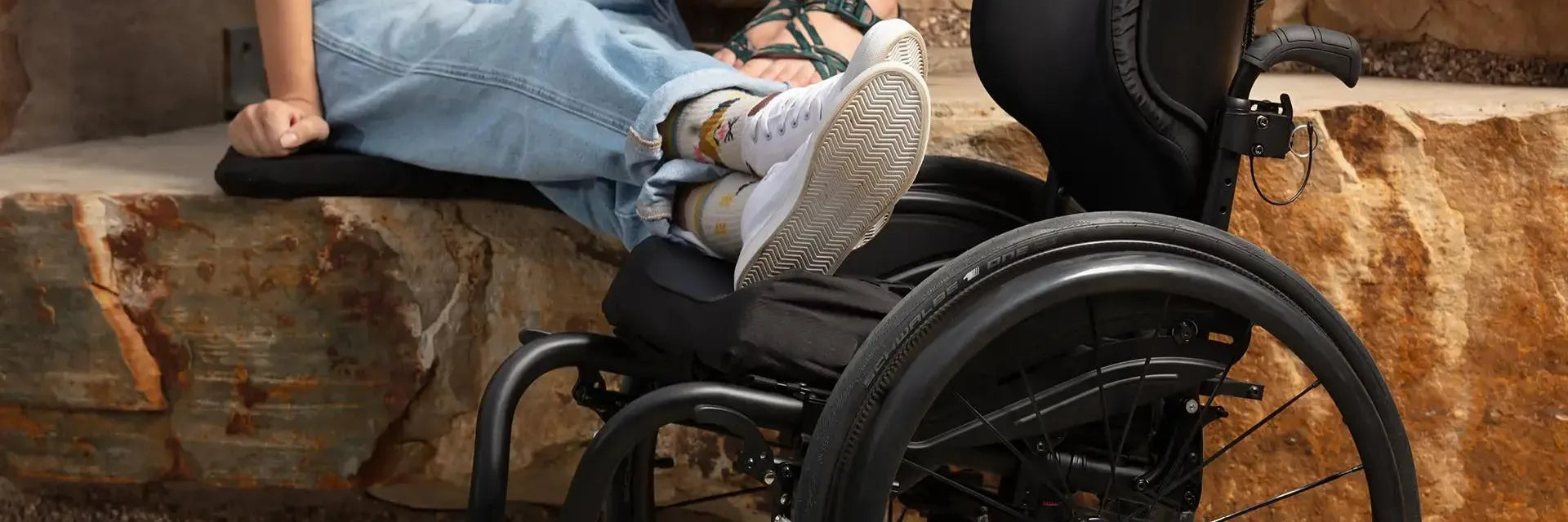A person sitting beside a black manual wheelchair with a contoured seat cushion and a shaped back support