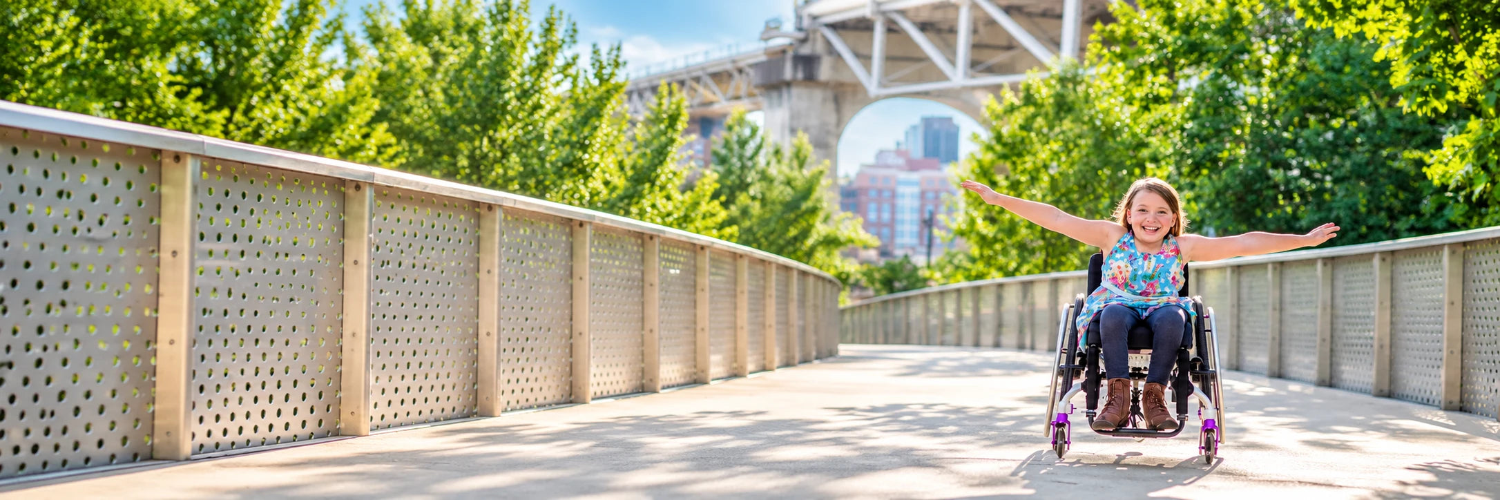 A smiling girl in a wheelchair with her arms spread wide on a riverside walkway under a bridge