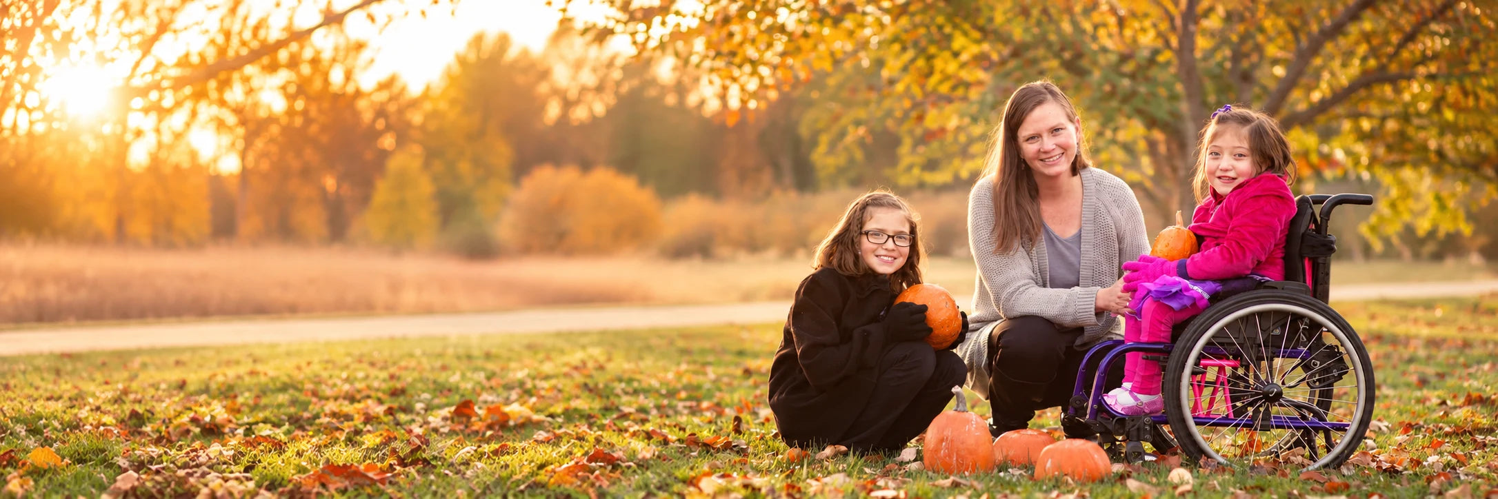 A mother kneeling in autumn leaves with her two daughters and their pumpkins, one girl seated in a pink and purple wheelchair