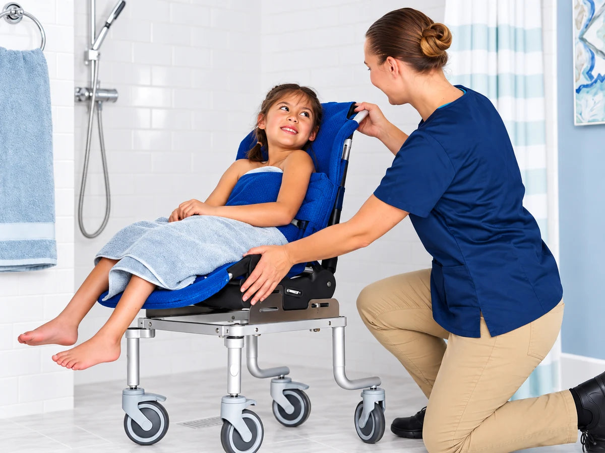 A caregiver kneeling beside a smiling girl wrapped in a towel on a reclined blue bath chair