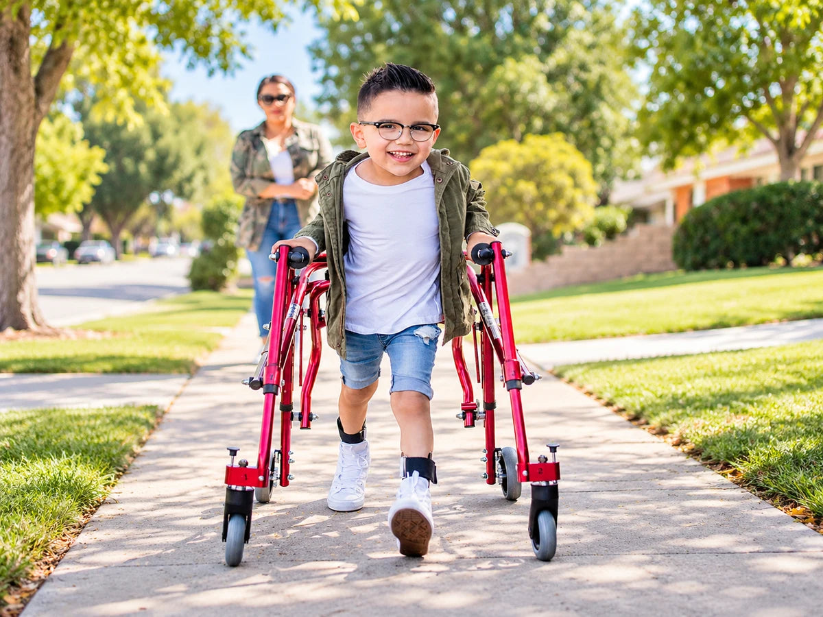A smiling boy walking down a sidewalk with a red walker while his mother watches