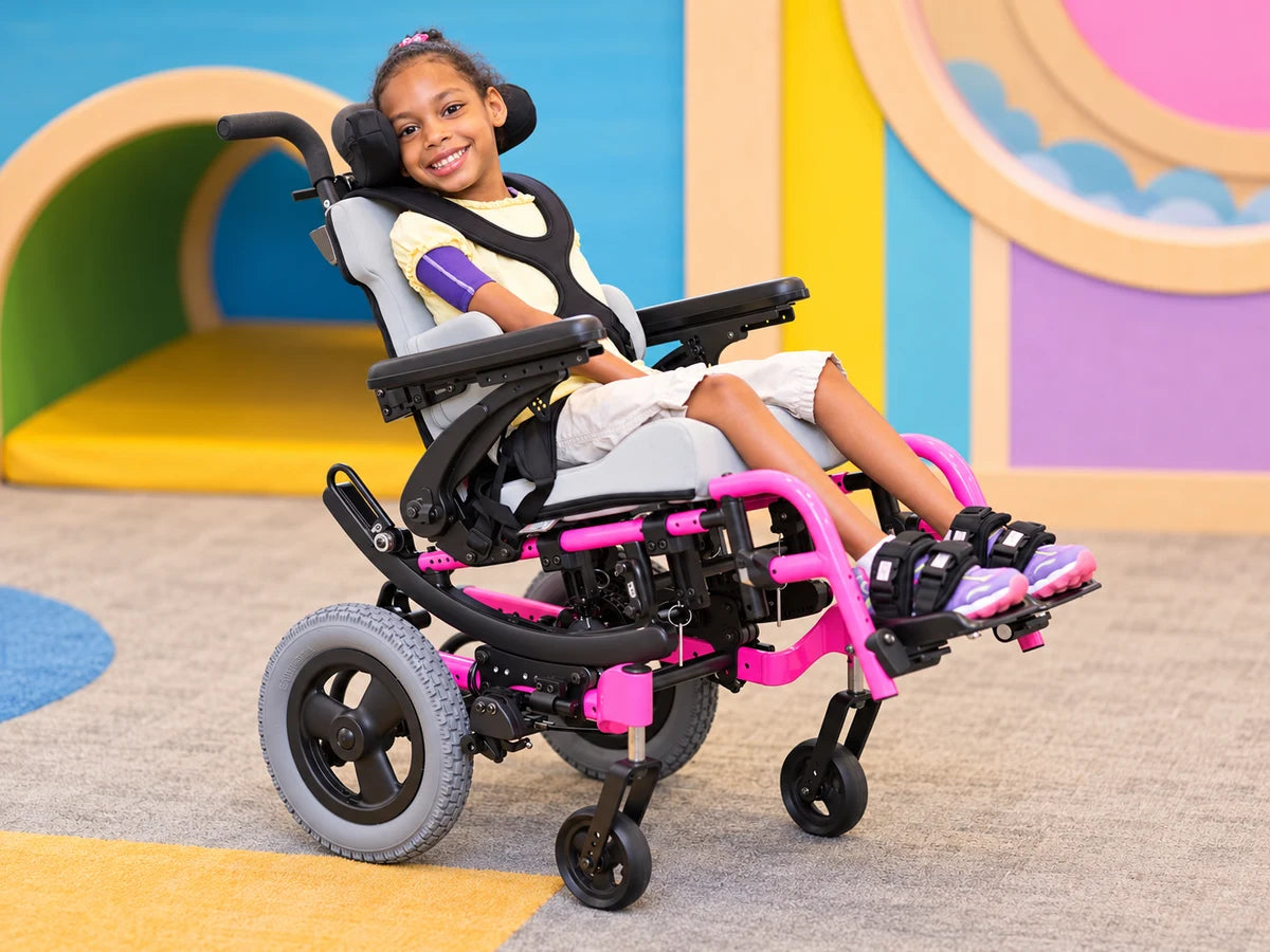 A smiling girl in a pink tilt in space wheelchair with a headrest and chest harness in a colorful playroom