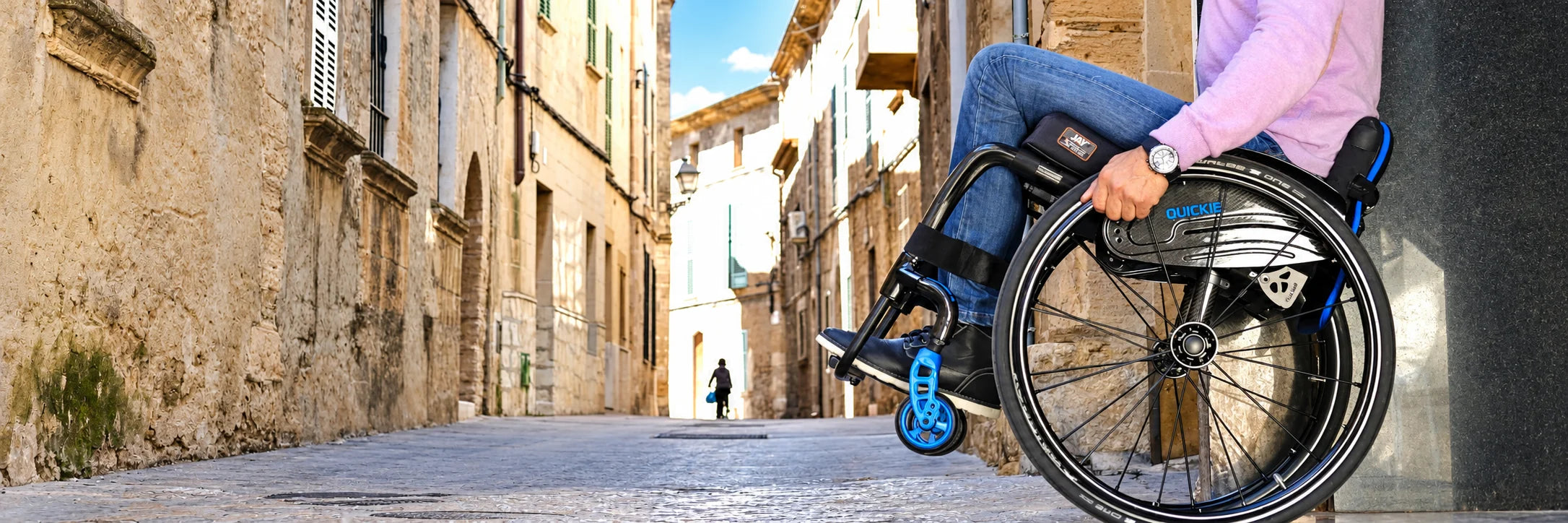 A man rolling himself down a stone street in a black ultralight manual wheelchair with blue casters