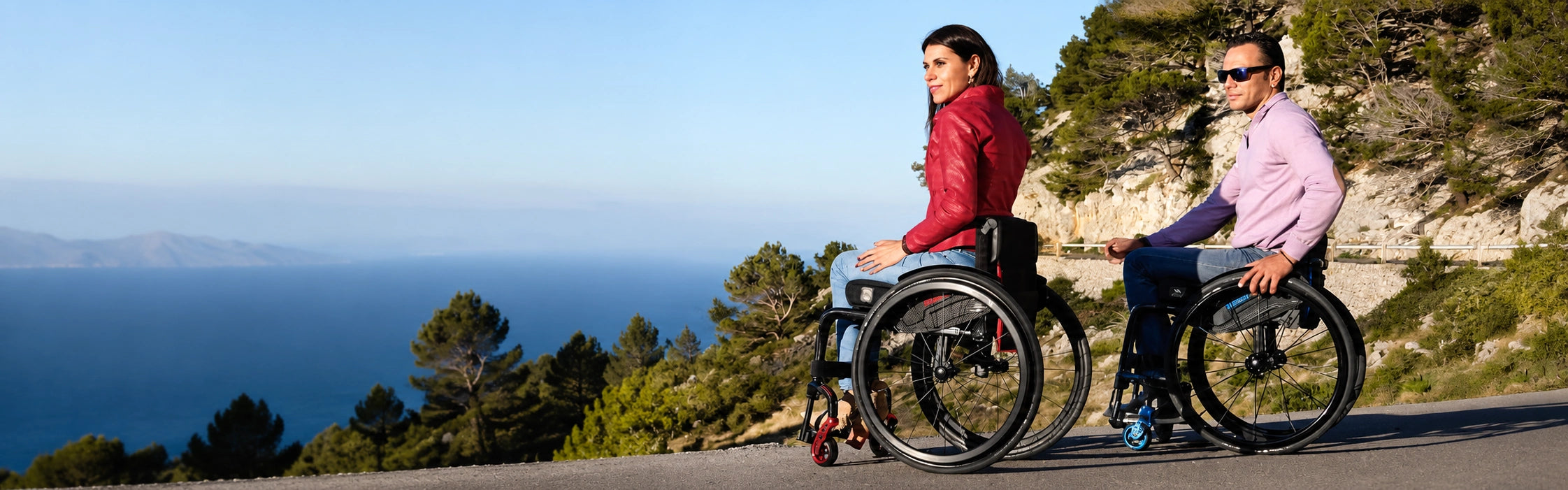 A man and a woman in ultra lightweight wheelchairs on a coastal road above the sea