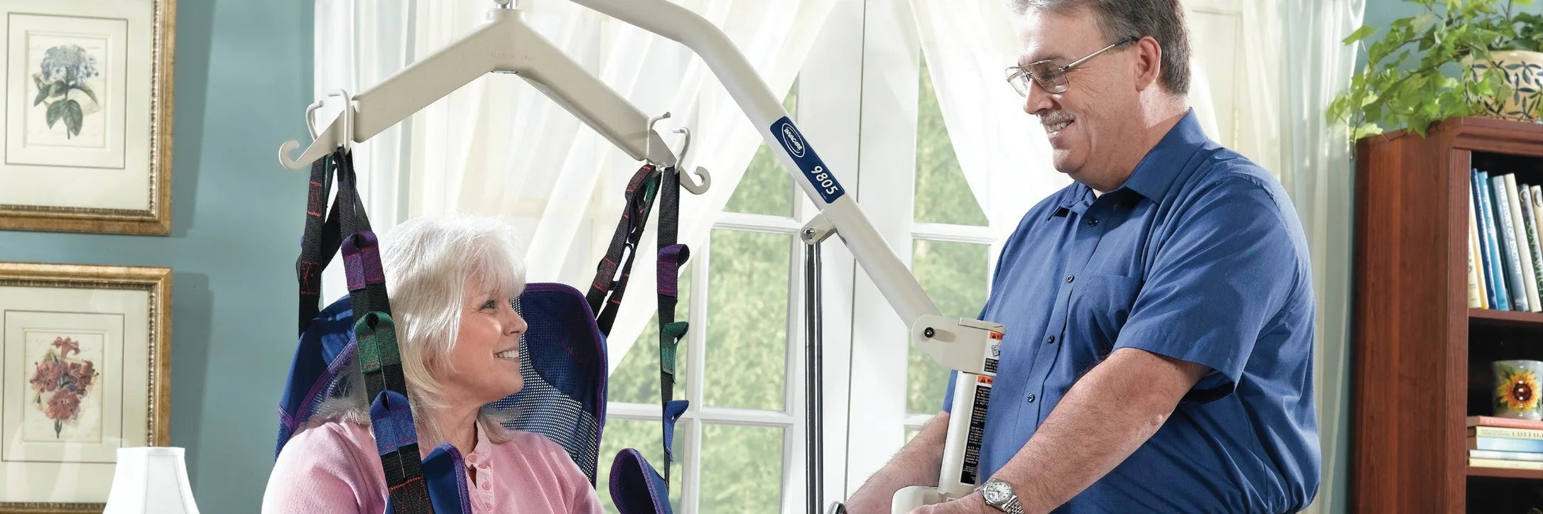 A smiling older woman raised in a full body sling on an electric lift while her husband works the hand control