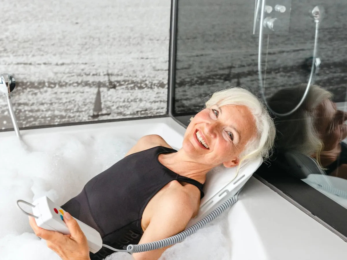 A smiling woman lying back on a bath lift in a bathtub, holding its remote