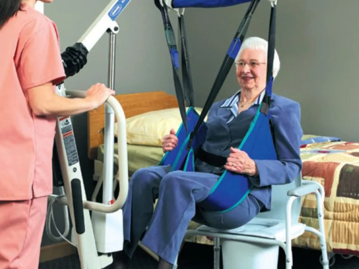 A woman seated in a blue toileting sling hung from a patient lift, beside a caregiver