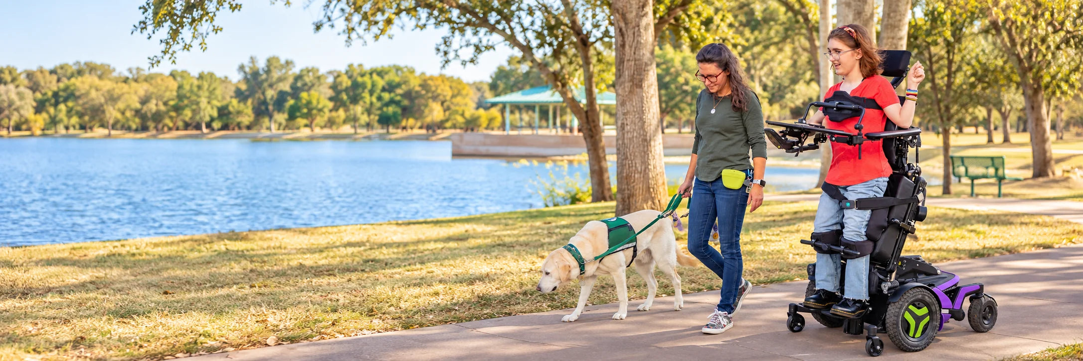 A young woman in a power standing wheelchair on a lakeside path in a park, beside a woman walking a service dog