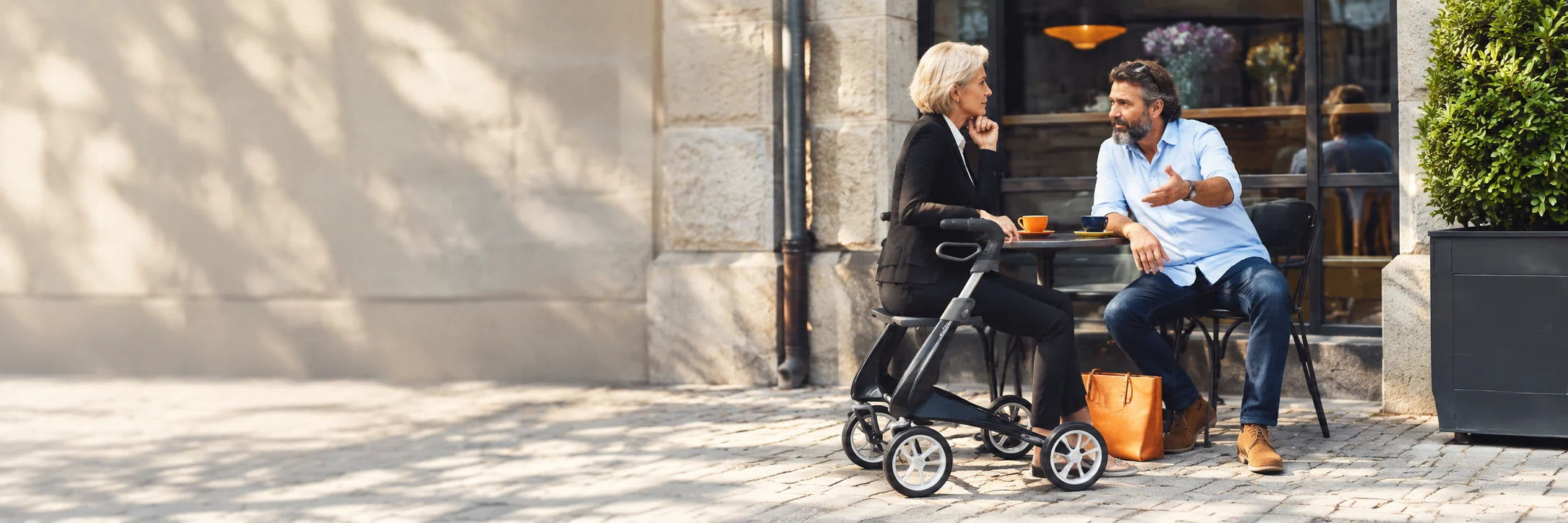 A woman sitting on her black rollator at a cafe table, talking with a man over coffee