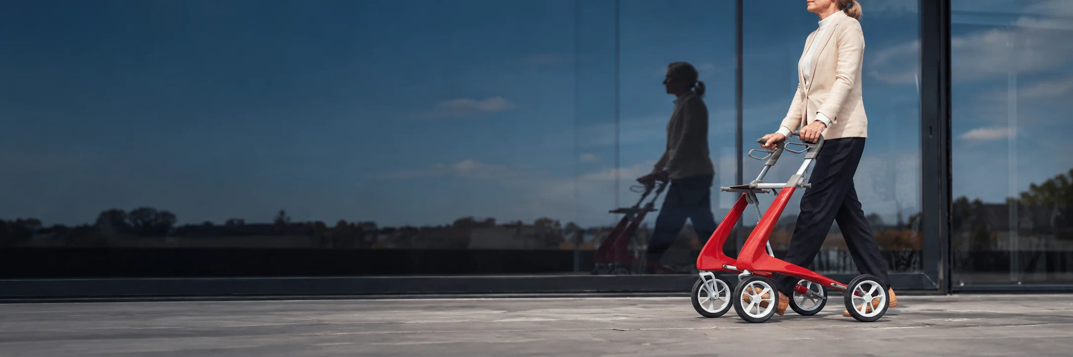 A woman walking with a red rollator past a glass wall that reflects the sky